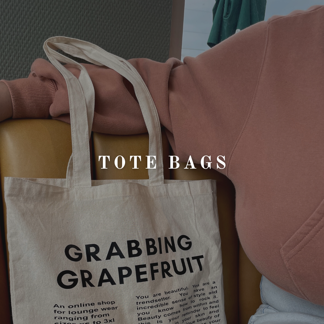 A shoulder and arm holds the straps of the Grabbing Grapefruit tote bag. In a coffee shop, the timeless tote bag displays the brand logo and an explanation of what Grabbing Grapefruit stands for and the meaning behind the fashion brand