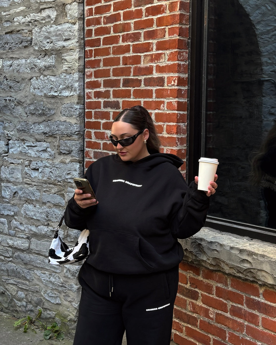Person in the Twilight and Tonic black hoodie and sweatpants using phone and holding a cup against a brick wall.