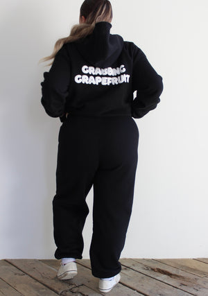 Person wearing a black hoodie with 'Grazing Grapefruit' text on the back, standing against a white wall.